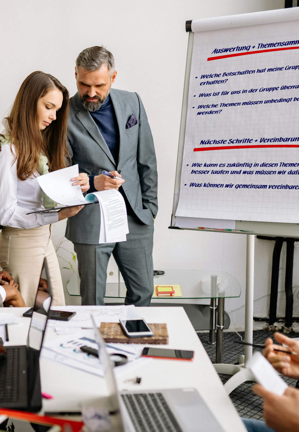 Zwei Mitarbeiter stehen neben einem Flipchart und blättern in Unterlagen.
